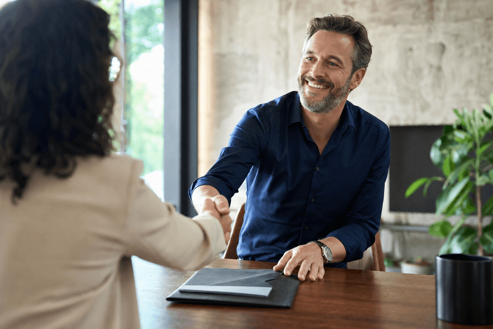 An agency owner shaking hands with a new client across a meeting table with a welcome folder.