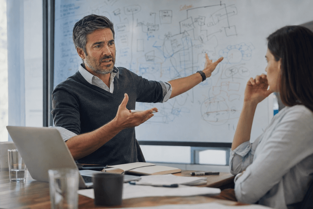 A consultant at a whiteboard covered in tangled diagrams, mid-explanation to a confused client.
